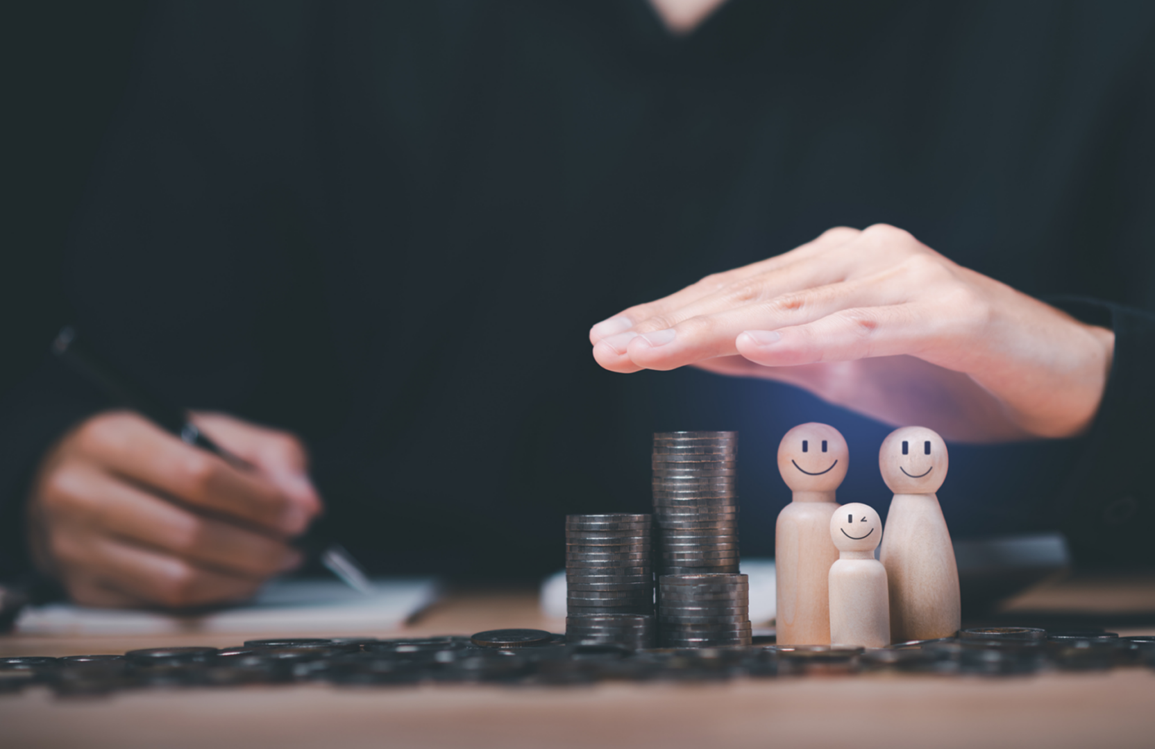 A hand shields wooden figures of a family next to stacked coins, symbolizing financial protection or insurance.